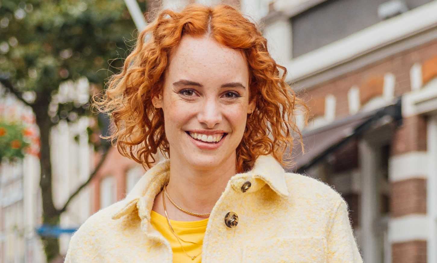 Dame met geel shirt staat voor een gebouw